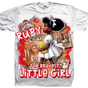 May include: White t-shirt featuring a graphic of Ruby Bridges, a young Black girl, walking with a book. The design includes the text "RUBY Bridges" and "THE BRAVEST LITTLE GIRL" in red and white.