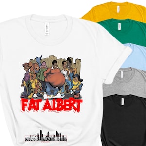 Op de afbeelding: Wit T-shirt met een cartoonafbeelding van Fat Albert en zijn vrienden, met de tekst "FAT ALBERT" in het rood. Andere T-shirts in verschillende kleuren zijn zichtbaar. Het shirt heeft een ronde hals en is gemaakt van een zacht materiaal.