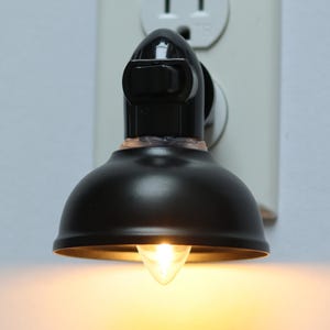 May include: A black plug-in night light with a round, dome-shaped shade and a built-in on/off switch. The light is plugged into a standard electrical outlet and is turned on, illuminating the room with a warm, yellow glow.