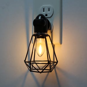 May include: A black plug-in night light with a diamond-shaped wire cage and a warm white light bulb.