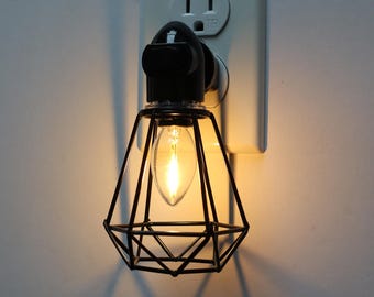 Könnte beinhalten: Ein schwarzes Steckdosen-Nachtlicht mit einem diamantförmigen Drahtkäfig und einer warmweißen Glühbirne.