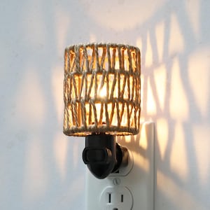 Op de afbeelding: Een wit stopcontact met een zwarte stekker voor een nachtlampje. Het nachtlampje heeft een geweven, beige, cilindrische kap.