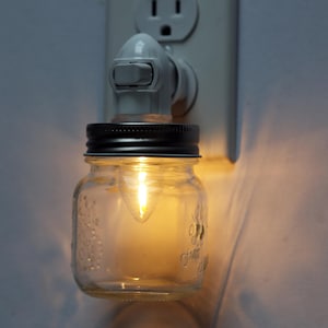 Puede incluir: Una luz nocturna blanca enchufable con un frasco de vidrio transparente y una tapa negra. La luz está encendida e ilumina el frasco.