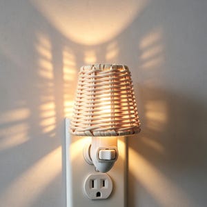 May include: A woven wicker night light with a conical shade. The night light is plugged into a white electrical outlet. The light is on, casting a warm glow and shadows on the wall. The night light is made of natural-coloured wicker.