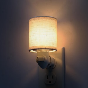 Op de afbeelding: Een klein, wit nachtlampje dat in een stopcontact is gestoken. Het nachtlampje heeft een cilindrische lampenkap van lichtgekleurde, getextureerde stof. De lamp is verlicht en geeft een warme gloed.