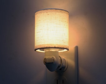 Op de afbeelding: Een klein, wit nachtlampje dat in een stopcontact is gestoken. Het nachtlampje heeft een cilindrische lampenkap van lichtgekleurde, getextureerde stof. De lamp is verlicht en geeft een warme gloed.