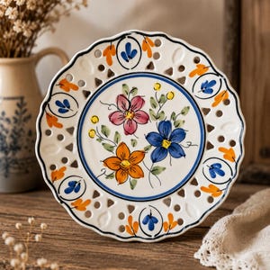 Może przedstawiać: Biały ceramiczny talerz z falistym brzegiem i dekoracyjnymi wycięciami. Talerz posiada centralny wzór z trzech kolorowych kwiatów: różowego, niebieskiego i pomarańczowego, otoczonych zielonymi liśćmi. Obrzeże ma pomarańczowe i niebieskie akcenty kwiatowe.