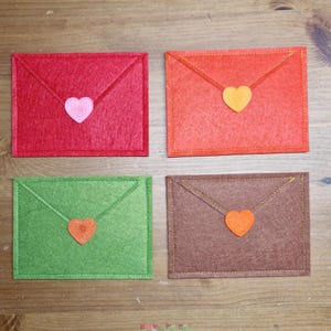 Può includere: Quattro buste di feltro colorate, rosse, arancioni, verdi e marroni, ciascuna con una chiusura a forma di cuore. Le buste sono disposte su una superficie di legno, mostrando un oggetto artigianale fatto a mano.