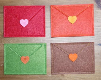 Può includere: Quattro buste di feltro colorate, rosse, arancioni, verdi e marroni, ciascuna con una chiusura a forma di cuore. Le buste sono disposte su una superficie di legno, mostrando un oggetto artigianale fatto a mano.