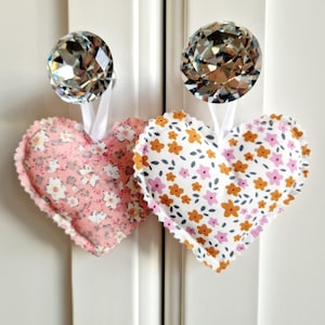 May include: Two fabric heart-shaped ornaments with floral patterns. The hearts are hanging from crystal-like knobs on a white cabinet door.