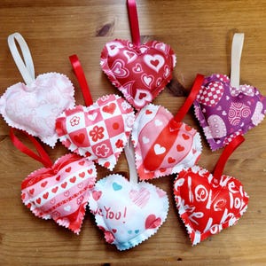 May include: Eight handmade felt heart ornaments with various Valentine's Day themed patterns. The hearts are pink, red, white, and purple with different patterns including hearts, stripes, polka dots, and checkered designs. Some of the hearts have the words "I love you" and "Love you!" printed on them.