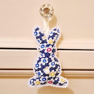 Puede incluir: Un conejito de tela azul y blanco con una cola de pompón blanco. El conejito está decorado con un patrón floral y tiene un borde de encaje blanco.
