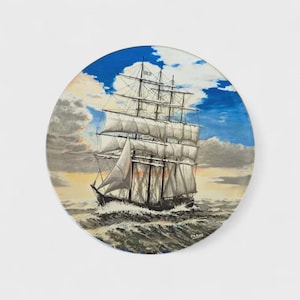 May include: Round coaster featuring a detailed painting of a tall ship sailing on a stormy sea. The artwork depicts the ship with billowing white sails against a blue sky with clouds. The coaster has a glossy finish.