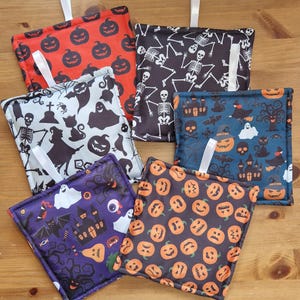 May include: Six square fabric items with Halloween patterns, each with a white ribbon loop. Designs include jack-o'-lanterns, skeletons, ghosts, haunted houses, and bats in orange, black, and purple. The items are square.