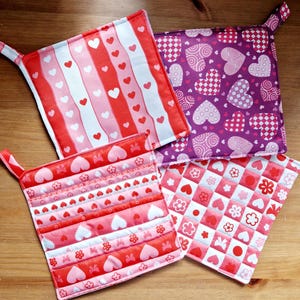 May include: Four quilted pot holders with a heart theme. The pot holders are in pink, red, white, and purple with different heart patterns.