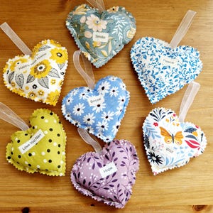Puede incluir: Ocho saquitos de tela en forma de corazón con diferentes patrones florales y mensajes bordados. Los mensajes incluyen "Happy New Home", "Smile", "Peace", "Bessie", "Love You Nan", "Wild", "Hope".