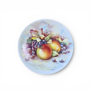 Puede incluir: Un plato decorativo con un bodegón de frutas pintadas. El diseño incluye un racimo de uvas moradas, dos manzanas y hojas verdes. El plato tiene un borde azul claro y un diseño de fruta pintada.