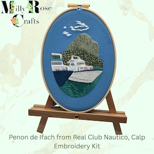 May include: An oval embroidery hoop with a blue fabric background features a detailed embroidered scene of a mountain, boats, and water. The text "Penon de Ifach from Real Club Nautico, Calp Embroidery Kit" is below the hoop. The hoop is displayed on a small wooden easel.