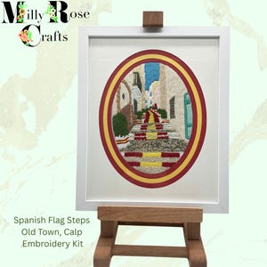 May include: Embroidered artwork of a Spanish street scene, framed in white. The image features a cobblestone street with steps, incorporating the colors of the Spanish flag. The artwork is displayed on a wooden easel. Text on the image reads: Spanish Flag Steps, Old Town, Calp, Embroidery Kit.