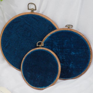 May include: Three round embroidery hoops with a teal blue velvet fabric stretched across the wooden frames. The hoops are nested inside each other, with the largest hoop at the top and the smallest hoop at the bottom.