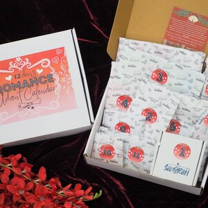 May include: A white box with a red and pink gradient background and the text "12 days of Romance Advent Calendar". The box is open and inside is a white box with 12 numbered envelopes. The envelopes are decorated with a snowflake pattern and the numbers 1 through 12 are visible. The box is sitting on a red velvet surface.