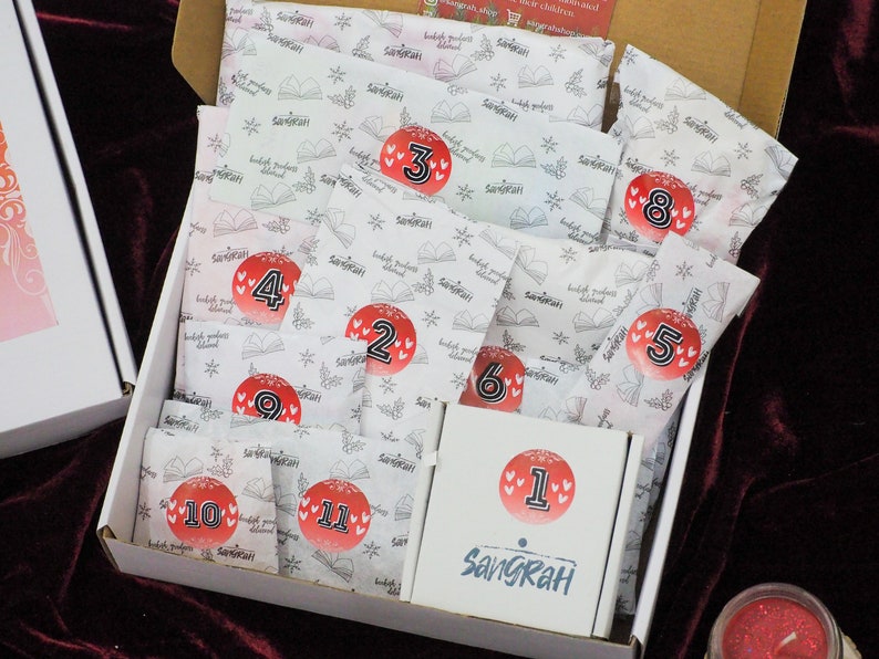 May include: A white box with a white and red pattern containing 11 numbered packages. The packages are labelled with numbers 1 through 11. The box is open and the packages are visible. The box is labelled "Sangrah".