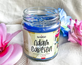 May include: A blue soy candle in a glass jar with a white label that reads "Sangrah Adam Carlsen White Musk - Cedarwood - Spice 5 oz. Soy Candle".