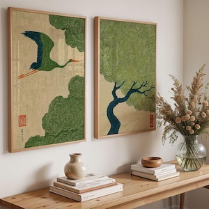 May include: Two framed rectangular wall art prints featuring a light beige background. The left print shows a green crane in flight with red accents and a red seal. The right print depicts a dark blue tree with a textured green canopy.