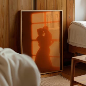 Könnte beinhalten: Ein rechteckiges gerahmtes Wandbild mit der leuchtend orangefarbenen Silhouette zweier sich umarmender Figuren. Das Bild zeigt ein Fenstergittermuster hinter den Figuren, das einen warmen, leuchtenden Effekt in einem einfachen hellen Holzrahmen erzeugt.