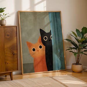 Può includere: Una stampa artistica incorniciata con due gatti dei cartoni animati. Uno è arancione, l'altro nero, entrambi con grandi occhi rotondi. Lo sfondo è verde e blu tenui, con una cornice in legno. L'opera d'arte è appoggiata a un muro.