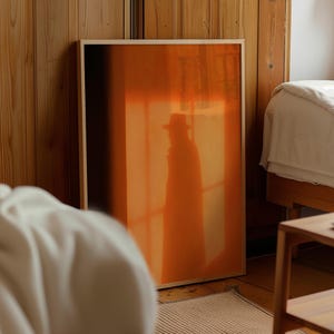 Könnte beinhalten: Ein rechteckiger gerahmter Wandkunstdruck mit einem lebendigen orangefarbenen und tiefschwarzen Farbverlauf. Im Mittelpunkt steht eine dunkle, schattenhafte Figur mit einem breitkrempigen Hut und einem langen Mantel vor einem warmen Hintergrund.
