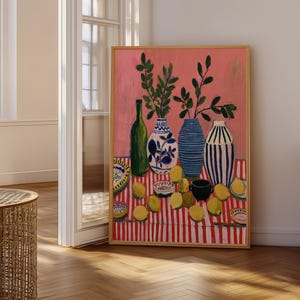 May include: A framed painting featuring a pink background with a table covered in a red and white striped cloth. The table holds a green bottle, three patterned vases with green leafy branches, several yellow lemons, and small decorative bowls.