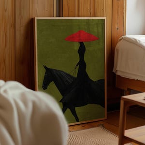 May include: A framed wall art print featuring a silhouette of a person in a long black dress and a wide red hat riding a black horse against a solid olive green background. The artwork is displayed in a light wood frame.