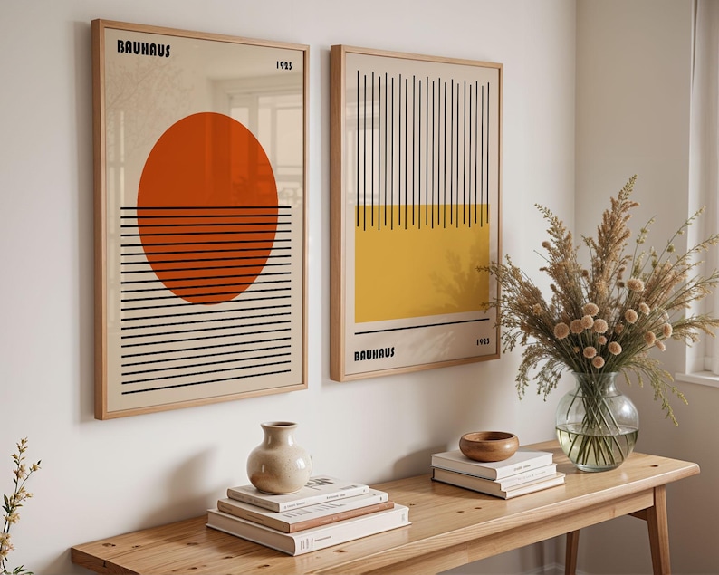a minimalist interior design with two framed abstract artworks hanging on a white wall above a wooden table. The artworks have a geometric design with bold lines and shapes, creating a modern and sophisticated aesthetic. On the table, there is a vase with flowers, a stack of books, and a bowl, adding a touch of warmth and personalization to the space.