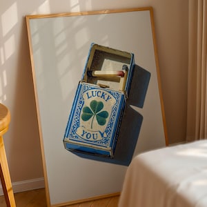 May include: A blue matchbox with the words "LUCKY YOU" and a shamrock design. The matchbox is open, revealing matches inside. The artwork is framed in a light wood frame.