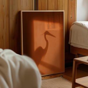 Könnte beinhalten: Ein rechteckiges gerahmtes Wandbild mit einem warmen, gebrannten orangefarbenen Hintergrund. Im Mittelpunkt steht die dunkle, deutliche Silhouette eines langhalsigen Vogels, der einem Reiher oder Kranich ähnelt, auf der orangefarbenen Fläche.