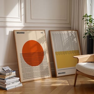 two framed artworks with abstract designs, placed on a wooden floor in a room with a chair and a stack of books.