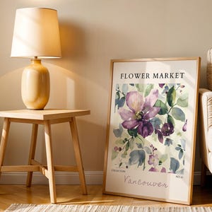 a wooden table with a lamp on top, a framed poster of a flower market, and a beige armchair in the background.