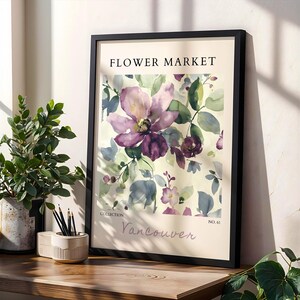 a framed poster of a watercolor painting of a purple flower, placed on a wooden shelf next to potted plants and a small white bowl.