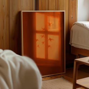 Peut inclure: Une impression encadrée rectangulaire présentant une image aux tons orange chaud d'un carreau de fenêtre. Le motif affiche la silhouette d'une volée d'oiseaux en plein vol sur un fond orange translucide et lumineux, dans un cadre en bois clair.