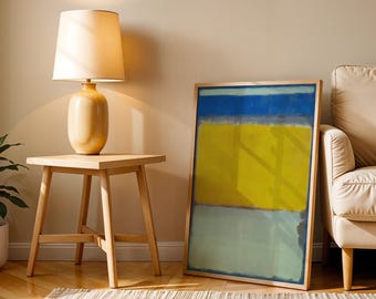Puede incluir: Una pieza de arte mural rectangular enmarcada que presenta bloques de color horizontales en azul profundo, amarillo vibrante y verde pálido. La obra se exhibe en un marco de madera clara y descansa en el suelo junto a una mesa auxiliar y una lámpara.