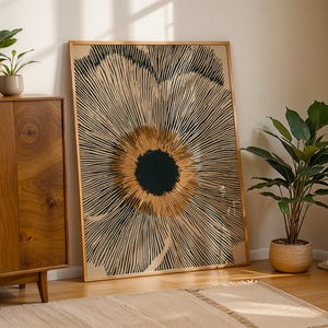 May include: A large rectangular framed wall art piece featuring a stylized, abstract floral design. The artwork displays a solid black circular center surrounded by radiating, thin black lines on a tan background, creating a sunburst effect.
