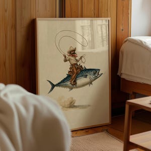 Pode incluir: Uma impressão de arte emoldurada apresentando um cowboy com chapéu bege, camisa branca e perneiras castanhas montado num grande atum azul e prateado. O cowboy segura um laço acima da cabeça e uma pequena nuvem de poeira surge sob o peixe.
