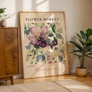 a large framed poster of a floral design, with the text "FLOWER MARKET" and "VANCOUVER" printed on it. The poster is displayed on a wooden cabinet, with a potted plant to the right and a rug on the floor in front of it.
