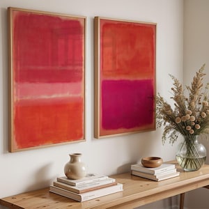 May include: Two framed abstract art prints with red and pink color blocks. The artwork is displayed above a wooden table with a vase of dried flowers, books, and decorative items. The frames are made of light-colored wood.