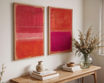 May include: Two framed abstract art prints with red and pink color blocks. The artwork is displayed above a wooden table with a vase of dried flowers, books, and decorative items. The frames are made of light-colored wood.