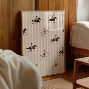 May include: A framed rectangular print featuring a repeating pattern of cowgirls riding horses and swinging lassos against a background of vertical light pink and white stripes. The artwork is encased in a simple light-colored wooden frame.