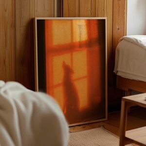 Könnte beinhalten: Ein rechteckiges gerahmtes Wandbild mit einer warmen, orangefarbenen Fotografie einer Hundesilhouette, die vor einem Fenster sitzt. Das Bild zeigt den Schatten des Tieres vor einem leuchtenden, sonnendurchfluteten Hintergrund mit sichtbaren Fensterscheiben.
