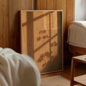 May include: A rectangular framed print featuring a warm, golden-toned surface with a dark, crisp shadow of a plant cast across it. The shadow displays intricate leaf patterns and stems, overlaid by the geometric shadow of a window frame.