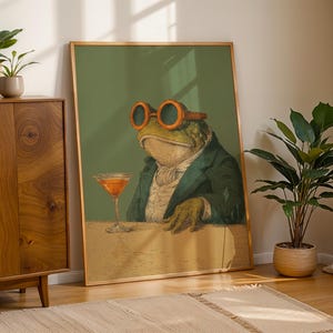 May include: A framed art print featuring a dapper frog wearing orange-rimmed glasses and a suit, holding a cocktail. The artwork has a vintage aesthetic with a green background and a tan table. The print is leaning against a wall.